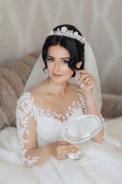 Uzun dantelli kolları ve şık bir tacı olan beyaz, seksi bir gelinlik giymiş esmer bir gelin odasında gri bir kanepede otururken aynaya bakıyor. Harika makyaj. Omuzlarını aç. Güzel eller. Uzun peçe. Gelinin sabahı.