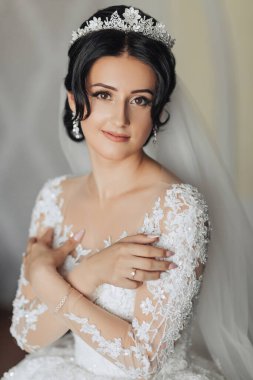 Uzun dantel kollu, uzun kollu, esmer bir gelin odasında gri bir kanepede otururken şık bir taç pozu veriyor. Harika makyaj. Omuzlarını aç. Güzel eller. Uzun peçe. Gelinin sabahı. Portre