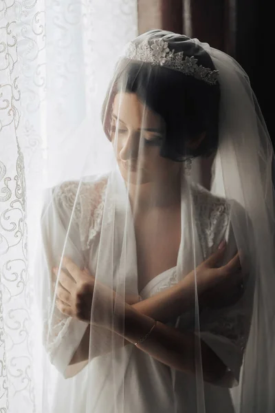 一个黑发新娘裹着面纱站在窗边朝下看 漂亮的妆容和头发厚的面纱 婚纱照漂亮的新娘 — 图库照片