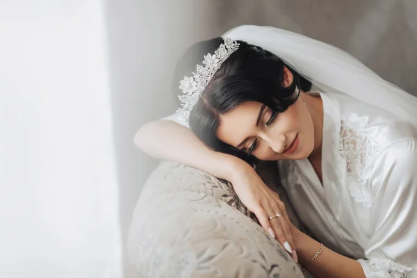一个穿着长袍 头戴时髦皇冠的黑发新娘躺在沙发的后面 眼睛紧闭着 漂亮的妆容和头发厚的面纱 婚纱照漂亮的新娘 — 图库照片