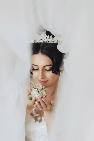 黑发新娘站在一个时髦的皇冠和白色的衣服 裹着面纱 向下看去 嗅着新郎的袖口 漂亮的妆容和头发厚的面纱 婚纱照漂亮的新娘 — 图库照片