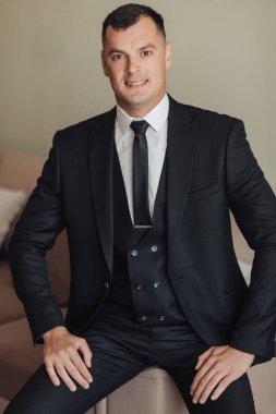 Beyaz gömlekli, siyah kravatlı ve siyah takım elbiseli bir adam koltukta otururken poz veriyor. Ön manzara. Şık bir saat. Erkek tarzı. Moda. İş dünyası