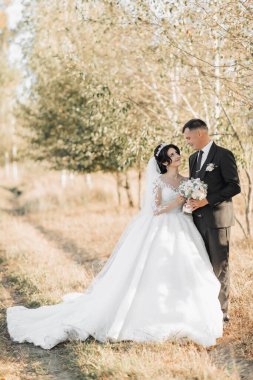 Doğada düğün fotoğrafı. Siyah elbiseli damat ve trenli beyaz elbiseli gelin ağaçların arka planında bir tarlada duruyorlar, gelin ve damat birbirlerine bakıyorlar ve bir buket tutuyorlar. Portre. Parlak yaz düğünü