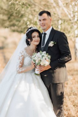 Doğada düğün fotoğrafı. Siyah takım elbiseli damat ve trenli beyaz elbiseli gelin ağaçların arkasında bir tarlada duruyorlar, gelin ve damat kameraya bakıyor. Portre. Parlak yaz düğünü