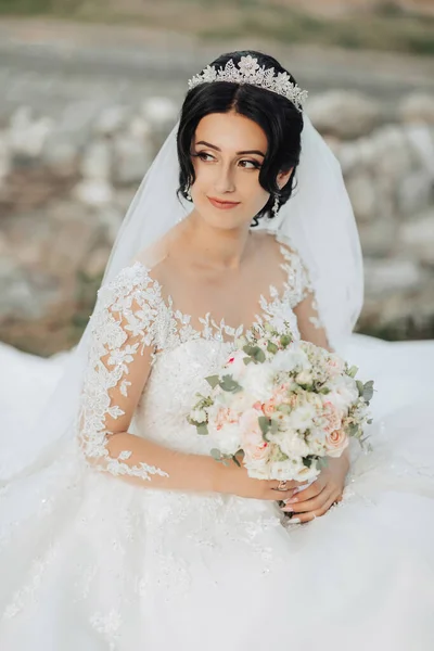 Portrait of the bride in nature. A brunette bride with a crown on her head, in a white voluminous dress sits, poses, holding a bouquet in her hands. Beautiful hair and makeup. Wedding shooting