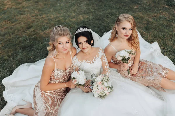 stock image A brunette bride in a white elegant dress with a crown and her blonde friends in gold dresses pose with bouquets while sitting on the grass. Wedding portrait in nature, wedding photo in a light tone. Three beautiful women