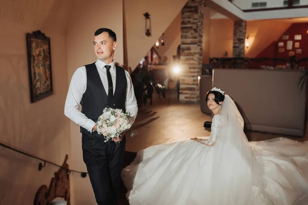 新娘和新郎的婚纱照 室内摄影 光线优美 一个穿着黑色背心的时髦新郎站在新娘面前 手里拿着一束鲜花 新娘坐在新娘身后 — 图库照片