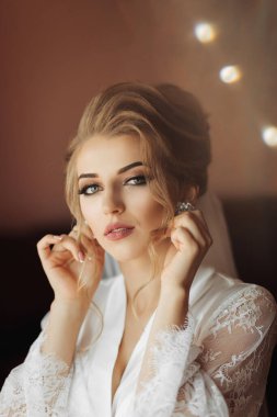 Uzun, dantelli etekli sarışın bir gelin odasında dikiliyor, pencerenin yanında poz veriyor, küpelerini takıyor. Güzel saç ve makyaj, açık büfe. Hafif bir tonda düğün portresi