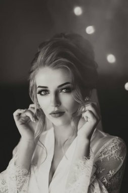 Siyah beyaz fotoğraf. Uzun, dantelli etekli sarışın bir gelin odasında dikiliyor, aynanın karşısında duruyor, küpelerini takıyor. Güzel saç ve makyaj, açık büfe. Düğün portresi. Aynadaki yansıma.