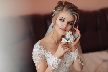 Uzun kollu dantel elbiseli sarışın gelin odasındaki deri koltukta oturmuş, poz veriyor ve küpe takıyor. Güzel saçlar, makyaj, açık omuzlar. Düğün portresi. Fransız manikürü.