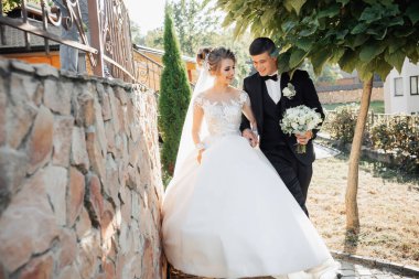 Düğün portresi. Siyah takım elbiseli damat ve sarışın gelin ağacın altındaki taş duvarın yanında durur damat gelinin elini öper. Doğada fotoğraf çekimi. Güzel saç ve makyaj.