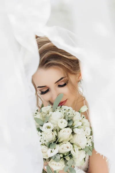 ベールで覆われた白いドレスに白いブロンドの花嫁は バラの花束でカメラのポーズをとっています 花嫁の肖像画 美しいメイクや髪 自然界の結婚式 — ストック写真