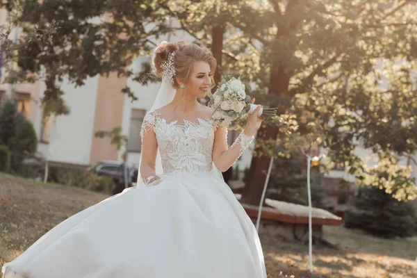 Wedding portrait. A blonde bride in a white dress with a train is walking, smiling and holding a bouquet and her wedding dress. Photo session in nature. Sun rays in the photo