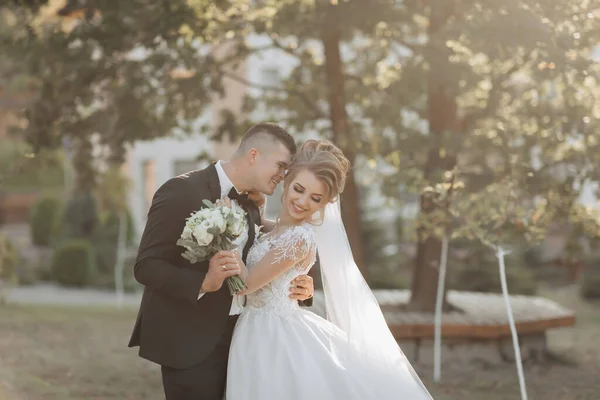 結婚式の肖像画 黒いスーツを着た新郎とブロンドの花嫁が立っていて 抱きついていて 木の下に花束を持っている 自然の中での撮影会 美しい髪とメイク — ストック写真