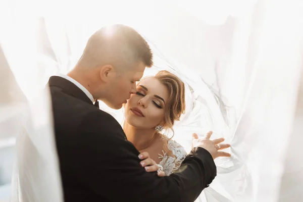 結婚式の肖像画 黒のスーツとブロンドの花嫁の新郎は 花嫁のベールに包まれて抱き合っている 空気中の長いドレス 自然の中での撮影会 美しい髪とメイク — ストック写真