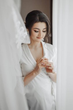 Portre. Peignoir giymiş esmer bir gelin, pencerede duruyor, alyansını tutuyor ve aşağıya bakıyor. Harika bir makyaj ve saç. Gönüllü duvak. Düğün fotoğrafı. Güzel gelin.
