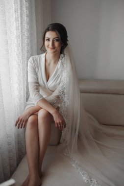 Gelinin portresi. Esmer bir gelin, çıplak bir kanepede bornozla ve uzun bir dantel peçeyle poz veriyor. Harika bir makyaj ve saç. Düğün fotoğrafı. Güzel gelin.