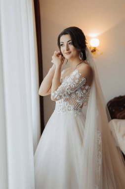 Uzun dantelli kolları olan, beyaz ve seksi elbiseli esmer bir gelin, küpelerini takıyor, odasında poz veriyor. Harika makyaj. Omuzlarını aç. Güzel eller. Uzun peçe. Gelinin sabahı. Portre