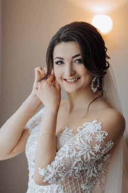 Uzun dantelli kolları olan, beyaz ve seksi elbiseli esmer bir gelin, küpelerini takıyor, odasında poz veriyor. Harika makyaj. Omuzlarını aç. Güzel eller. Uzun peçe. Gelinin sabahı. Portre