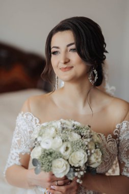 Beyaz elbiseli esmer bir gelin düğün buketini tutar ve poz verir. Fransız manikürü. Omuzlarını aç. Güzel eller. Uzun peçe. Gelinin sabahı. Ayrıntılar