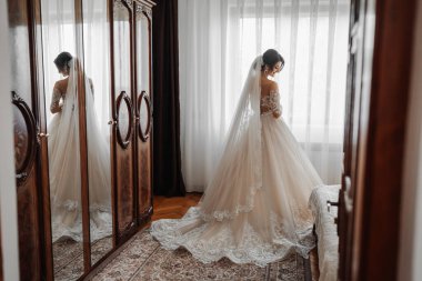 Beyaz elbiseli esmer bir gelin ve uzun bir duvak pencerenin önünde duruyor. Güzel eller. Fransız manikürü. Güzel saç ve makyaj. Gün ışığı