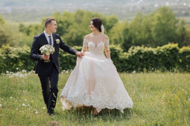 Gelin ve damadın doğadaki portresi. Uzun dantel elbiseli şık bir gelin ve damat bahçede el ele tutuşuyorlar. Birbirine aşık çift