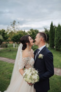 Gelin ve damadın doğadaki portresi. Uzun dantelli bir elbise içinde şık bir gelin ve damat bahçede ağaçların yanında sarılıp öpüşüyor. Aşık mutlu bir çift.