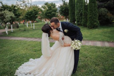 Gelin ve damadın doğadaki portresi. Uzun dantelli bir elbise içinde şık bir gelin ve damat bahçede ağaçların yanında sarılıp öpüşüyor. Aşık mutlu bir çift.