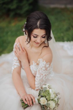 Doğadaki gelinin portresi. Beyaz dantelli gelinlik giymiş esmer bir gelin çimlerin üzerinde oturuyor, elinde buketle poz veriyor. Güzel saç ve makyaj. Omuzlarını aç. Düğün çekimi.