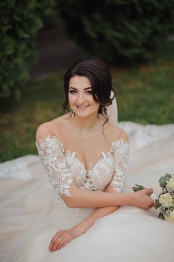Doğadaki gelinin portresi. Beyaz dantelli gelinlik giymiş esmer bir gelin çimlerin üzerinde oturuyor, elinde buketle poz veriyor. Güzel saç ve makyaj. Omuzlarını aç. Düğün çekimi.