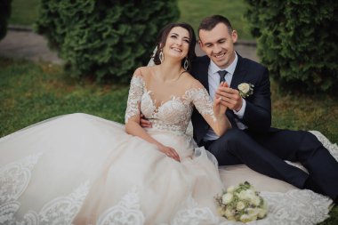 Doğadaki gelinin portresi. Beyaz dantelli bir gelinlik içinde esmer bir gelin ve siyah takım elbiseli şık bir damat çimlerin üzerinde oturmuş, poz veriyor, elinde buket tutuyorlar. Güzel makyaj. Omuzlarını aç.