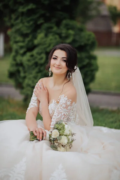 自然の中で花嫁の肖像画 白いレースのドレスを着たブルネットの花嫁が花束を持って ポーズをとって芝生の上に座っています 美しい髪とメイク 肩を開いて ウェディング撮影 — ストック写真