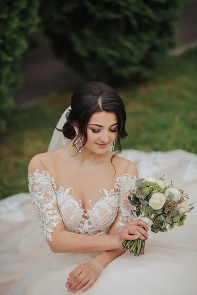 自然の中で花嫁の肖像画 白いレースのドレスを着たブルネットの花嫁が花束を持って ポーズをとって芝生の上に座っています 美しい髪とメイク 肩を開いて ウェディング撮影 — ストック写真