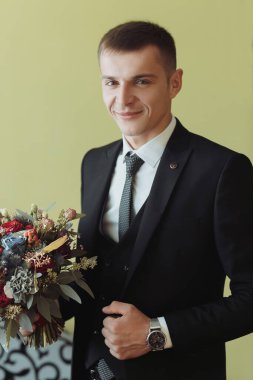 Portre. Beyaz gömlekli, siyah ceketli ve siyah takım elbiseli bir adam bir kanepede oturuyor, elinde farklı güllerden bir buket tutuyor. Şık bir saat. Erkek tarzı. Moda. İş dünyası