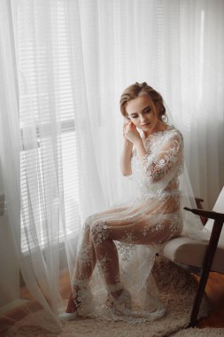 Şeffaf bornozlu gelin, parlak bir odada yumuşak pembe bir sandalyenin kenarına oturur ve zarif bir altın küpe takar.