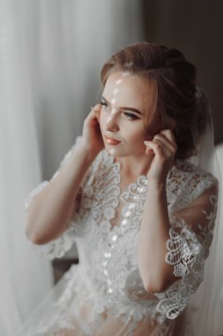 Şeffaf bornozlu gelin, parlak bir odada yumuşak pembe bir sandalyenin kenarına oturur ve zarif bir altın küpe takar.