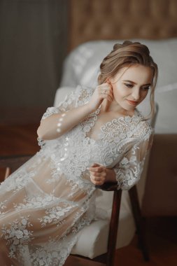 Şeffaf bornozlu gelin, parlak bir odada yumuşak pembe bir sandalyenin kenarına oturur ve zarif bir altın küpe takar.