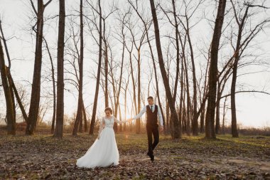 Düğün fotoğrafı. Gelin ve damat ormanda yürüyorlar. Damat sevgilisinin elini tutar. Uzun gelinlik. Uzun ağaçların arasında birbirine aşık bir çift. Sonbahar güneş ışığı.