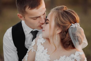 Doğada düğün fotoğrafı. Damat ahşap bir stantta oturuyor, gelin onun yanında duruyor, omuzlarına sarılıyor ve onu öpüyor. Gelin ve damadın portresi