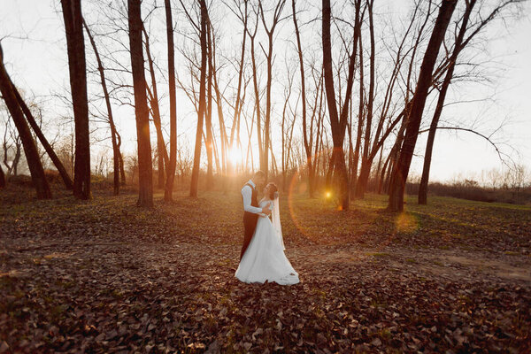 Wedding photo. The bride and groom are walking in the forest. The groom hugs the shoulders of his beloved. Long wedding dress. A couple in love among tall trees. Autumn sunlight.