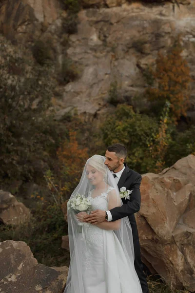 花嫁と新郎の岩の山の大きな石の横に高い上に立っている 花嫁はベールの下にあり 新郎は彼女の肩を抱いています — ストック写真