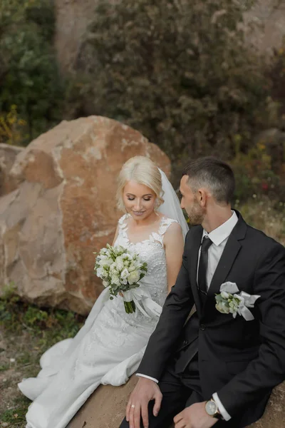 幸せな美しい花嫁とエレガントなスタイリッシュな新郎は岩の上に座って — ストック写真