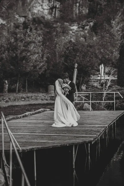 身穿黑色西服的新郎和穿着白色婚纱的新娘在码头上跳舞 在湖边和岩石旁边 黑白照片 — 图库照片
