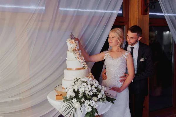 一个用鲜花和黄金装饰的白色婚礼蛋糕 矗立在一个玻璃架上 婚礼用的鲜花甜食 新娘和新郎在蛋糕旁边 — 图库照片