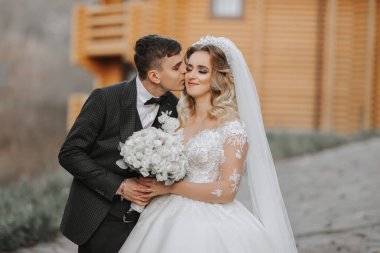 Parktaki modern ahşap evlerin yanında yakışıklı damat ve büyüleyici gelin bir arada. Portre. Uzun trenli muhteşem bir elbise. Zarif genç damat.
