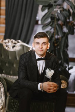 Düğün günü modern otel odasındaki koyu yeşil kanepede oturan ve gülümseyen siyah gömlekli pozitif genç damat portresi.