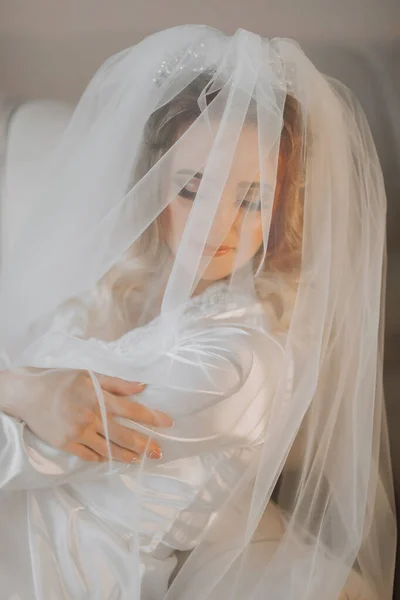 金发碧眼的新娘身穿缎袍 躺在沙发上 头戴面纱 美丽的头发和化妆 张开的胸围 婚礼肖像 面带微笑 — 图库照片