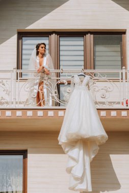 Balkon korkuluklarında asılı beyaz gelinlik, evin önünde geniş bir kare ve balkonda bir kız. Düğün detayları, uzun trenli modern gelinlik, uzun kollu ve arkası açık.