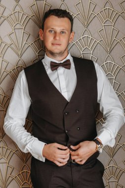 Zarif bir iş adamı camdan dışarı bakarken kahverengi bir yeleğin düğmelerini ilikliyor. Odadaki portre.