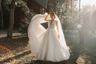 Düğün fotoğrafı. Gelin ve damat ormanda yürüyorlar. Damat bir buket tutuyor ve o da güzel bir şekilde uzun elbisesini kusuyor. Birbirine aşık bir çift. Yaz ışığı.